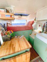 Cozy RV rental interior with a bed facing a stunning mountain view. Features sage green cabinets, a wooden table, and small kitchen.