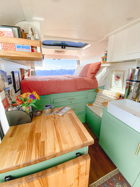 Cozy RV rental interior with a bed facing a stunning mountain view. Features sage green cabinets, a wooden table, and small kitchen.