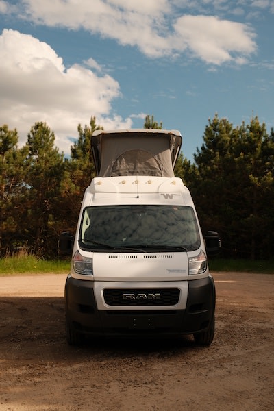 White pop-top Ram camper van on a dirt path with trees, perfect for your RV rental adventure.