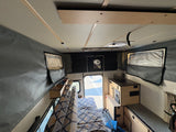 Cozy rv rental interior: patterned bench, compact kitchen, pop-top walls, and a view through the open back door.
