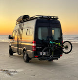 Black Mercedes Sprinter van, an ideal rv rental, on a golden sandy beach at sunset with a mountain bike. Van life adventure.