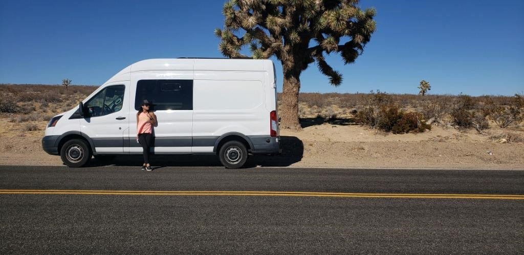 Desert adventure with an RV rental. A woman next to a white camper van on a sunny road, likely Joshua Tree NP.