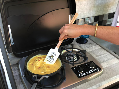 A person stirs eggs in a pan on a FlameKing stove with a bee spatula, making breakfast in an RV rental kitchen.