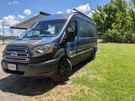 This customized Ford Transit camper van, likely an RV rental, features a unique livery, extended awning, and is parked in a grassy area.