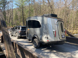 An RV rental, an Airstream Basecamp, towed by an SUV on a rustic bridge. Ready for outdoor RV adventures in nature.