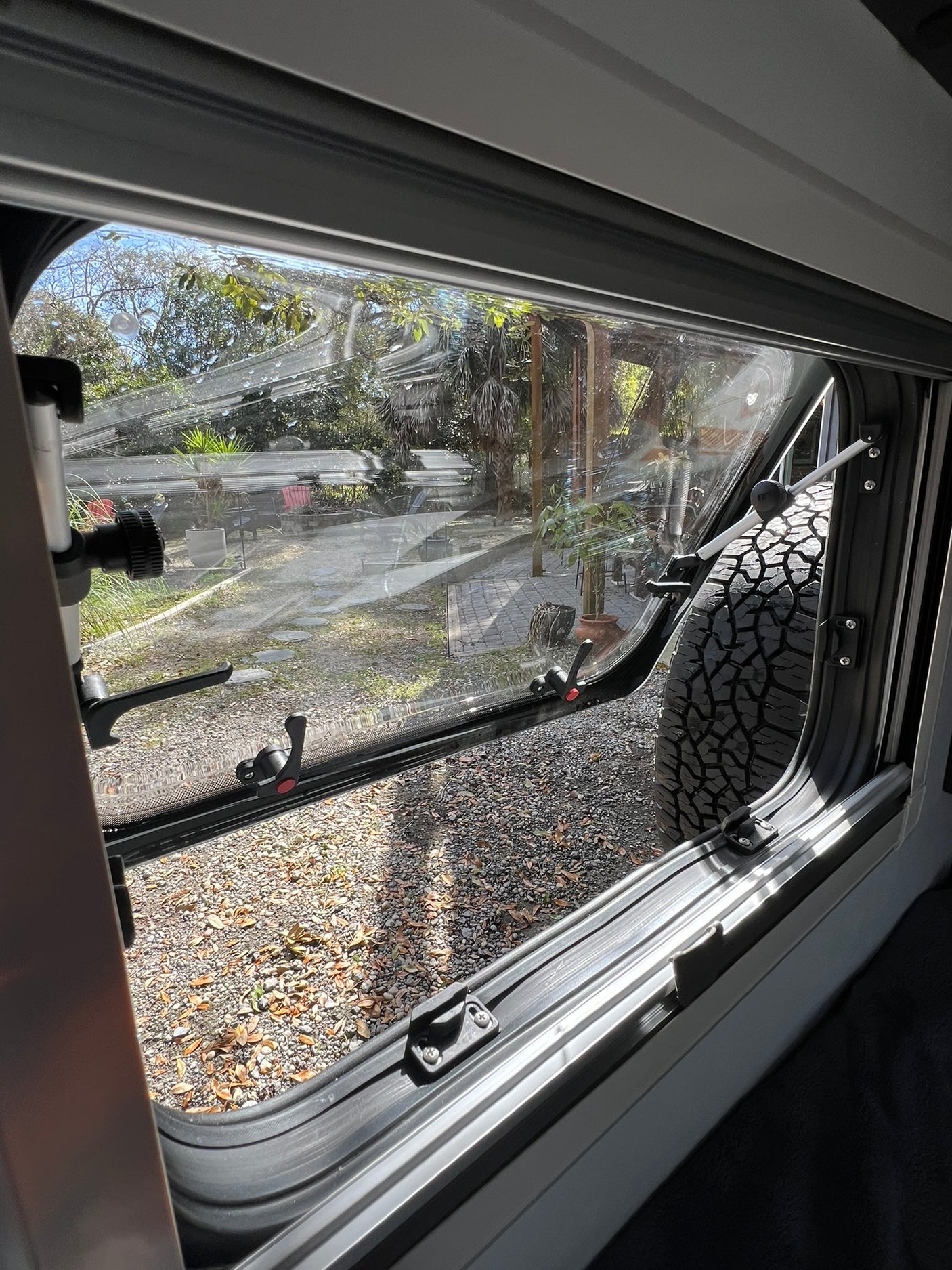 A partially open RV rental window offers a view of a gravel path, trees, and a large vehicle tire. Ready for adventure!