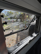 A partially open RV rental window offers a view of a gravel path, trees, and a large vehicle tire. Ready for adventure!