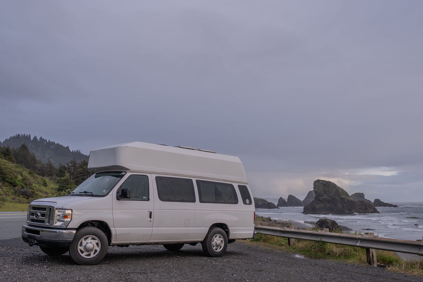 RV rental van parked on a gravel shoulder by an overcast coast with sea stacks and forested hills.