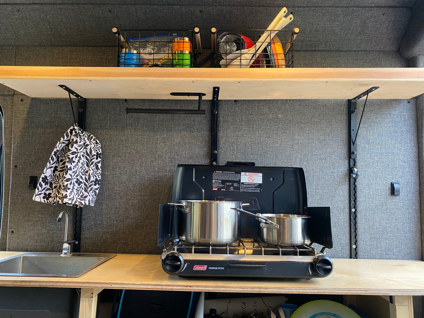 RV rental kitchen: compact setup with Coleman stove, pots, sink, & overhead storage. A handy towel hangs nearby. — photo 3