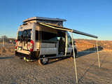 A silver RV rental van, with pop-top and awning extended, sits on a gravel lot under clear skies.