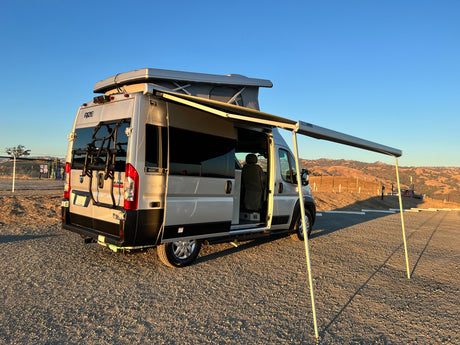 A silver RV rental van, with pop-top and awning extended, sits on a gravel lot under clear skies.