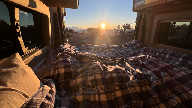 A cozy plaid bed inside an RV rental, offering a breathtaking sunrise view over rocky terrain and distant mountains.