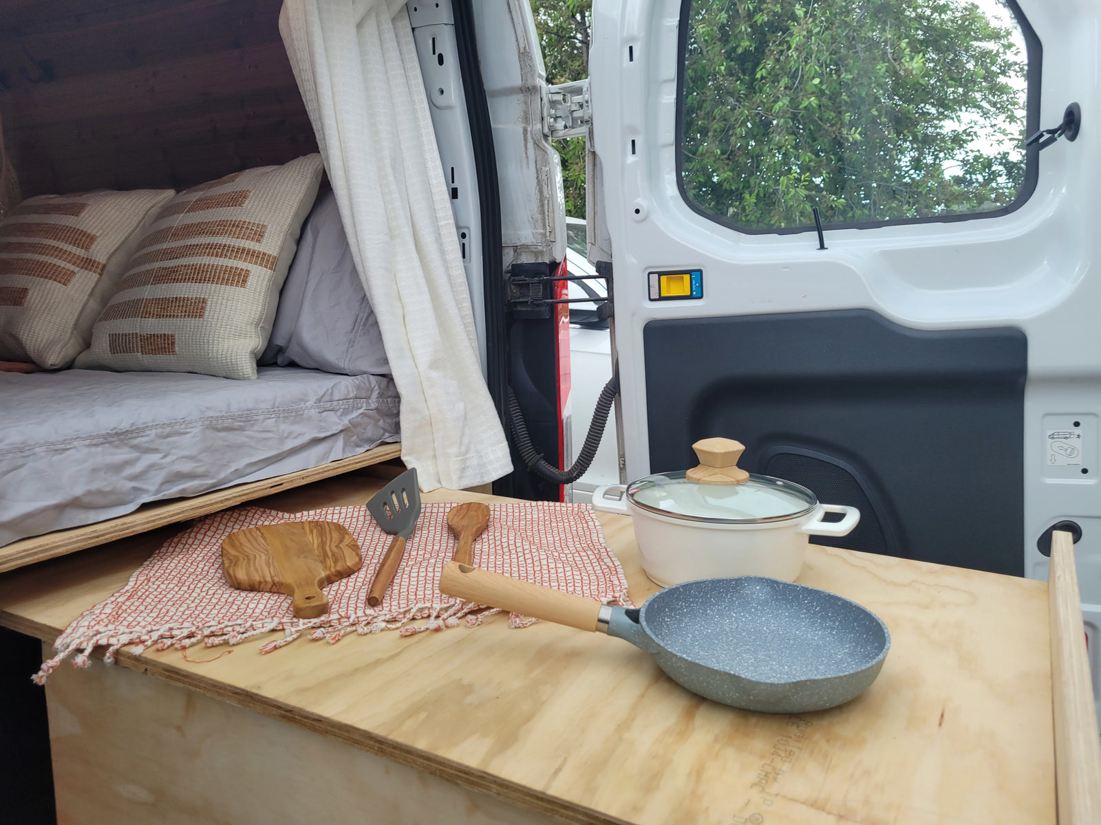RV rental interior showcasing a cozy bed and a practical kitchen setup with a pan, pot, and wooden utensils on a counter.