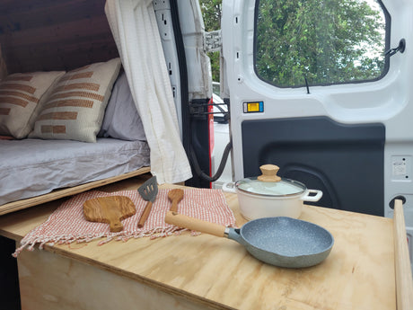 RV rental interior showcasing a cozy bed and a practical kitchen setup with a pan, pot, and wooden utensils on a counter.