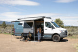 An rv rental camper van provides outdoor comfort for a family; parents and baby relax under an extended awning.