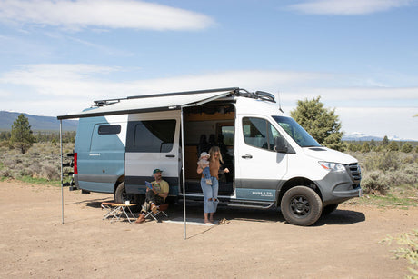 An rv rental camper van provides outdoor comfort for a family; parents and baby relax under an extended awning.