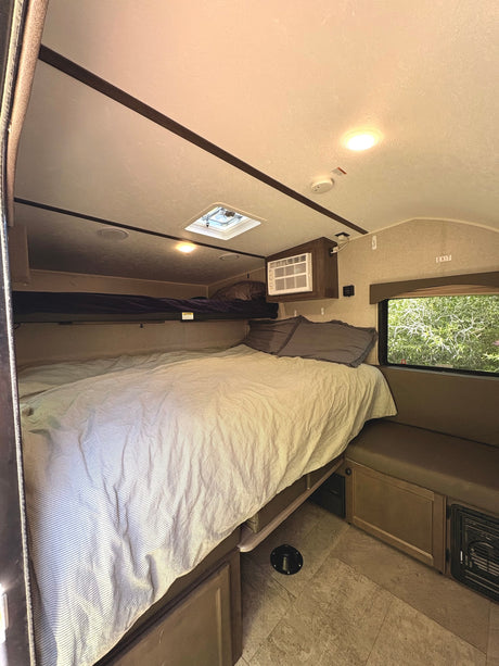 Cozy RV rental interior featuring a main bed with striped bedding, an upper bunk, and a side dinette by a window.