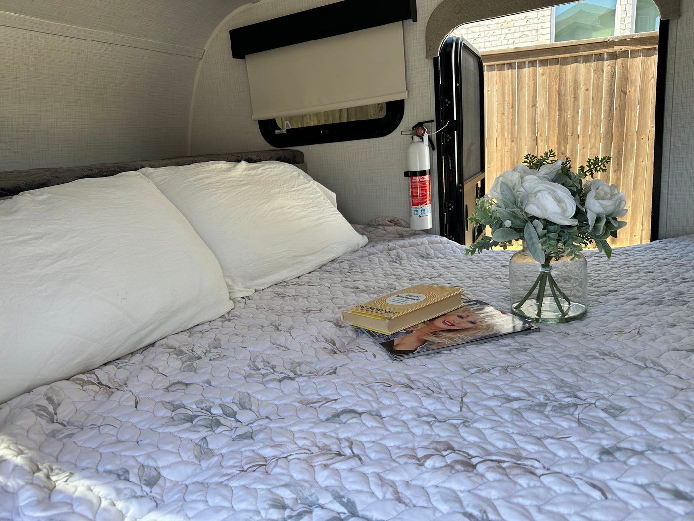 Cozy rv rental bedroom with a patterned quilt, white pillows, book, magazine, and flowers. Fire extinguisher visible near open door.