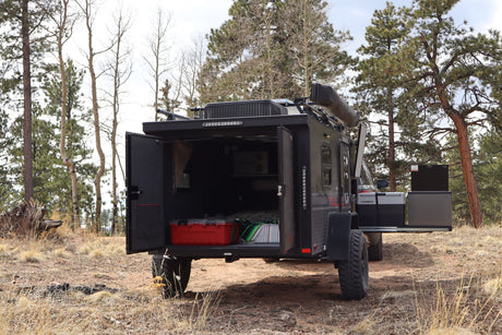 This rugged rv rental overland trailer is ready for adventure in the woods. Its rear hatch is open, revealing a bed, storage, and a Dometic fridge.