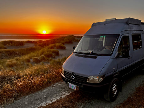 An RV rental camper van is parked by sandy dunes, offering a prime view of a stunning Oregon coast sunset over the ocean.