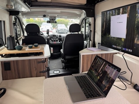 Inside an RV rental: a modern workspace with a laptop, monitor, & tea setup in the kitchen area, ideal for remote work.