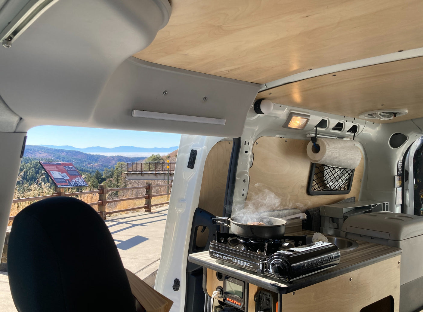 RV rental camper van with a portable stove cooking inside, offering a scenic mountain view from the open door.