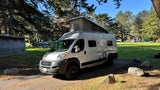 This rv rental, a white camper van with a pop-top, is parked at a sunny forested campground with other campers visible.