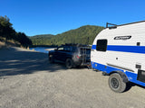 This SUV tows a white & blue Sunray Sport RV rental by a scenic river and forested mountains, ideal for outdoor adventures.