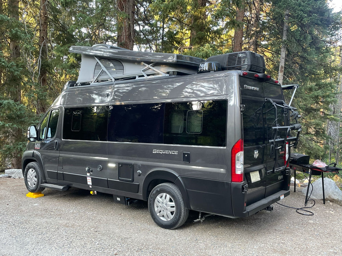 A gray pop-top camper van (RV rental) parked on gravel in a forest, with a grill set up for outdoor cooking. Ideal for camping travel. — photo 6