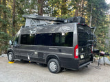 A gray pop-top camper van (RV rental) parked on gravel in a forest, with a grill set up for outdoor cooking. Ideal for camping travel.