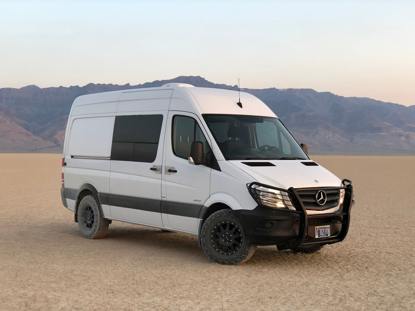 This white Mercedes-Benz Sprinter **RV rental**, fitted for off-road, rests on a desert lakebed with distant mountains. — photo 4