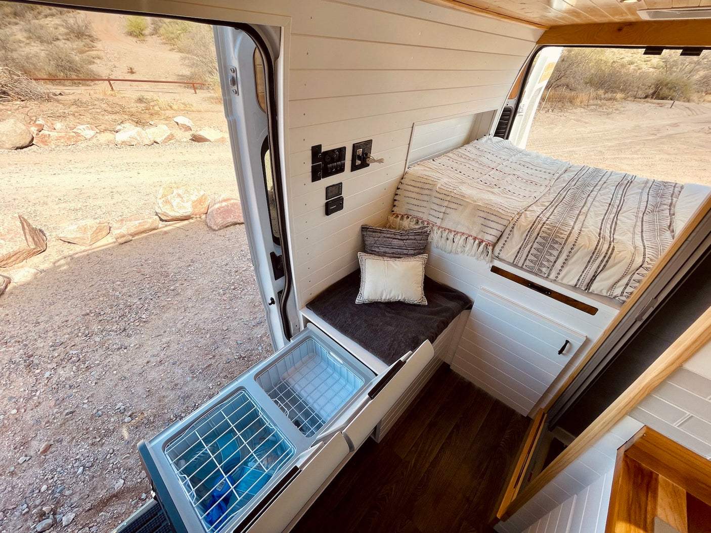 RV rental: Camper van interior showing bed, pull-out fridge, and controls, with a desert view from the open door.