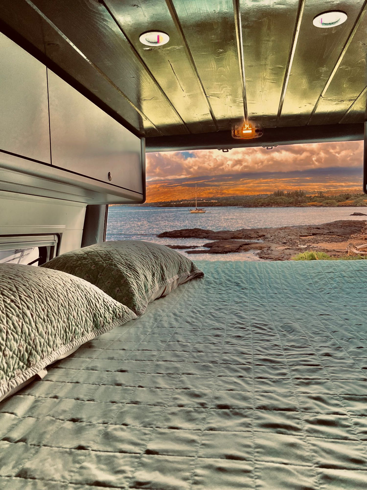 Wake up to paradise! This cozy RV rental features a comfortable bed overlooking a stunning ocean sunset with a distant sailboat.|Inside your **rv rental**: a cozy camper van kitchen with sink