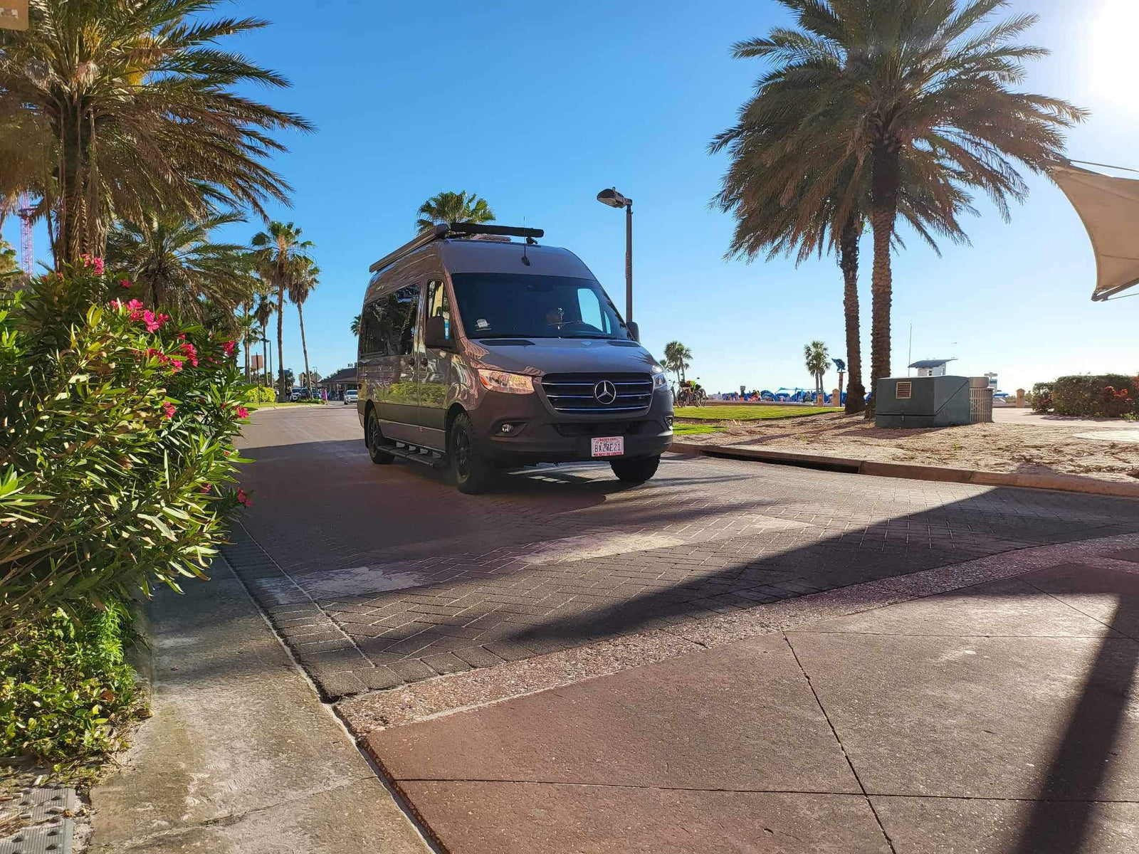 A dark grey Mercedes-Benz Sprinter RV rental drives on a sunny street lined with palm trees near the beach.