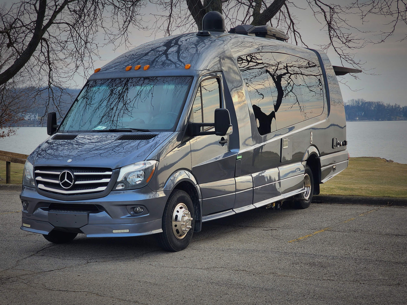 Luxury Mercedes-Benz Sprinter RV rental parked by a lake, reflections of bare trees on its shiny exterior.