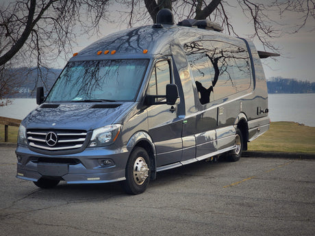 Luxury Mercedes-Benz Sprinter RV rental parked by a lake, reflections of bare trees on its shiny exterior.