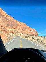 Drive your RV rental through stunning red rock country on a clear day. The road winds past towering cliffs with other cars ahead.
