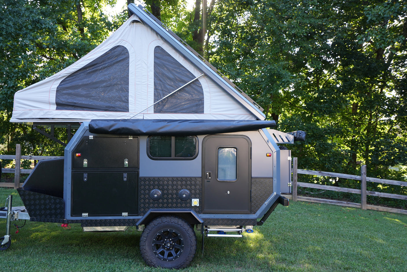 A rugged pop-top off-road camper trailer, perfect for an rv rental, stands on green grass with a forest backdrop.