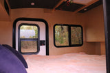 Cozy RV rental interior: a bed with pink blanket, wood walls, and windows looking out to autumn trees. Camper living.