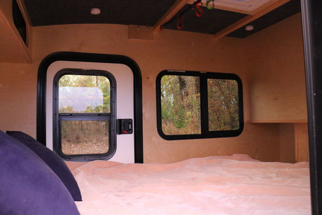 Cozy RV rental interior: a bed with pink blanket, wood walls, and windows looking out to autumn trees. Camper living.