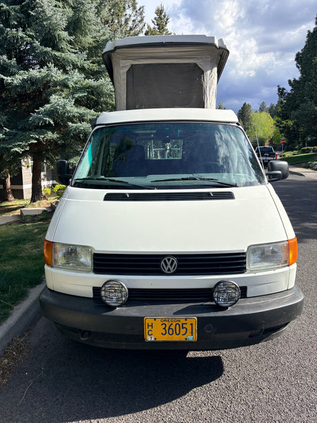 Front view of a white Volkswagen pop-top camper van, perhaps an RV rental, parked on a street by trees. Oregon plate visible.
