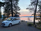RV rental van parked by a serene lake at sunset, surrounded by trees. A person relaxes on the shore. Perfect camping adventure.