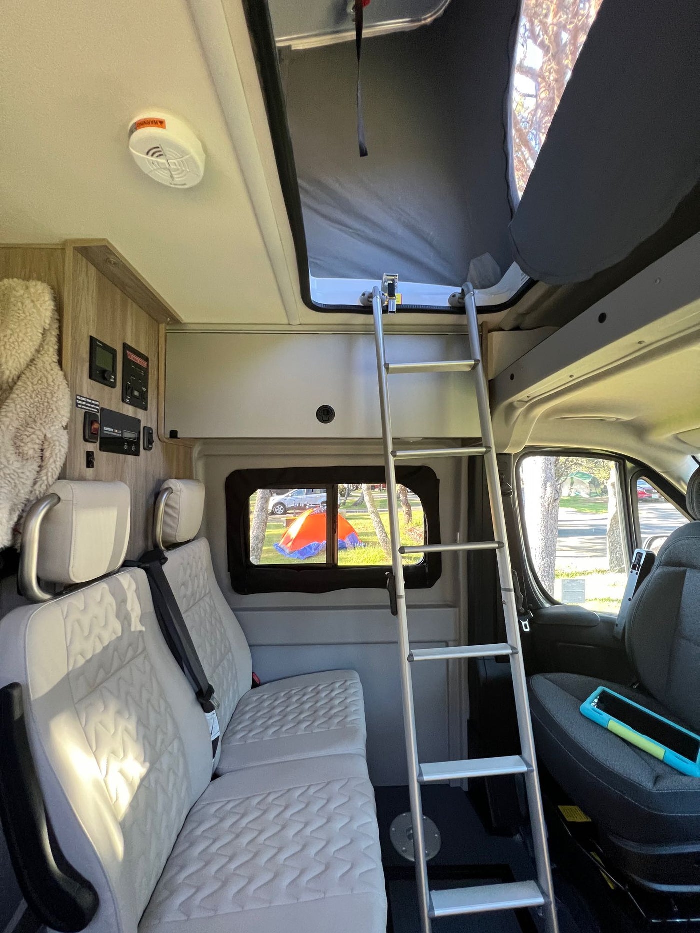  a person looks out the back window at a scenic view from their bed. Overhead cabinets are visible.|Inside an rv rental: a camper van's interior showing pop-top bed access via ladder — photo 9