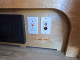 RV rental power panel: GFCI AC outlet, dual USB, 12V socket. Black electronic unit with green lights in camper.