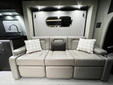 Modern RV rental interior: Light-colored segmented sofa, plaid and dark pillows, kitchenette, and sleek oval mirror on the wall.