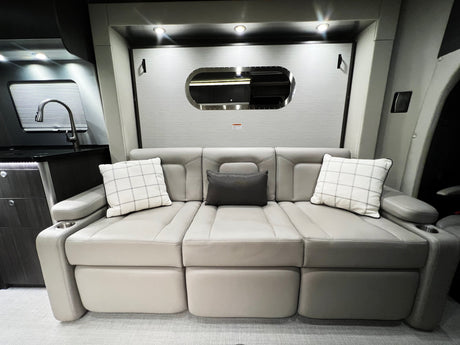 Modern RV rental interior: Light-colored segmented sofa, plaid and dark pillows, kitchenette, and sleek oval mirror on the wall.