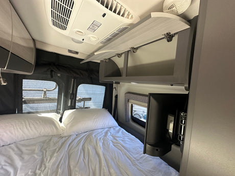 Inside an rv rental: a comfy white bed, ceiling AC, and open overhead cabinets for storage.