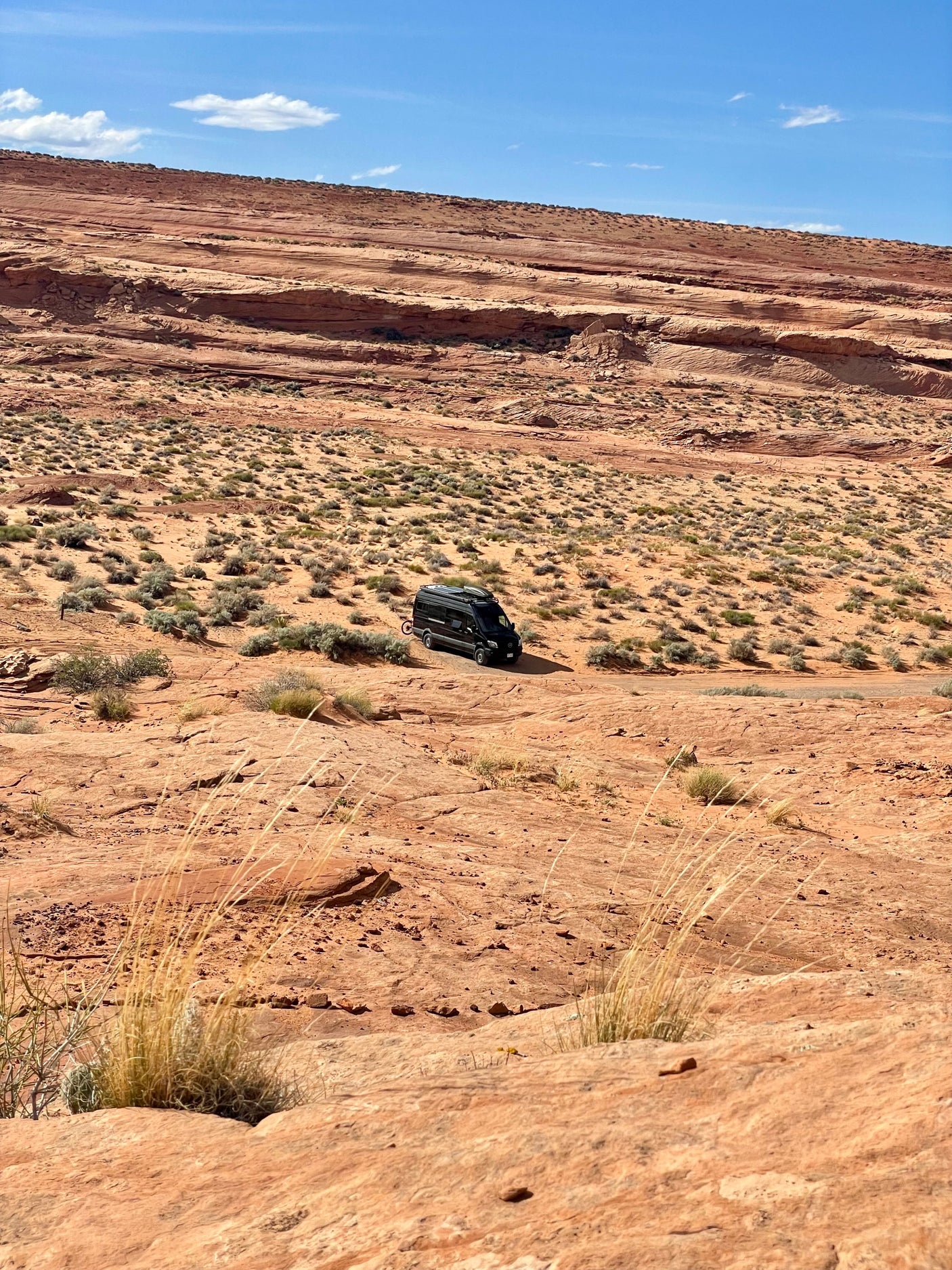 Explore the red rock desert in a black RV rental, parked amidst sparse brush under a bright blue sky.