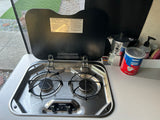 An rv rental's compact kitchen with a two-burner gas stove, instructions on lid, and coffee maker for travel cooking.