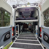 The rear of an RV rental van with open doors, revealing a raised bed, modular storage, and an extended loading ramp.